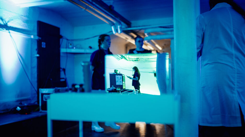 A photo taken a mirror of a nurse, with a blurred background containing other figures in a lab.